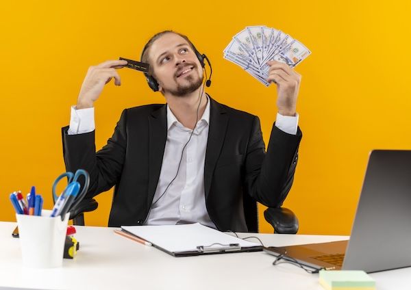 Elegante uomo d'affari in abito e cuffie con microfono imbottito e carta di credito, con sguardo sognante, seduto al tavolo in ufficio su sfondo arancione.
