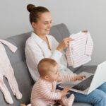 Inquadratura di una donna sorridente e soddisfatta con una camicia bianca e jeans, seduta sul divano con la madre di sua figlia, che mostra la camicia dei bambini allo schermo del portatile, esprimendo emozioni positive sullo shopping per abbigliamento online bambini