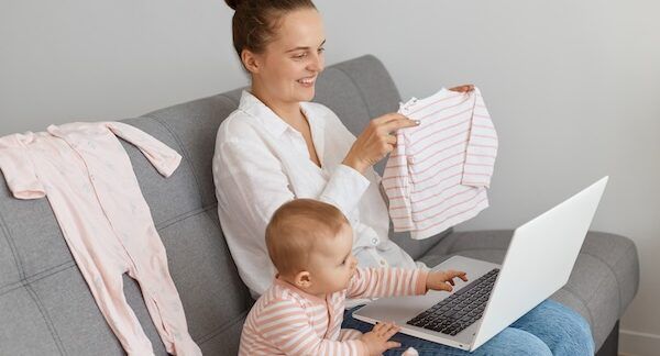 Inquadratura di una donna sorridente e soddisfatta con una camicia bianca e jeans, seduta sul divano con la madre di sua figlia, che mostra la camicia dei bambini allo schermo del portatile, esprimendo emozioni positive sullo shopping per abbigliamento online bambini