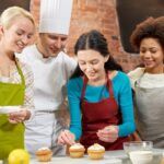 Concetto di scuole di cucina professionali, gastronomia, panificazione, cibo e persone - gruppo felice di donne e chef maschio che cucinano in cucina